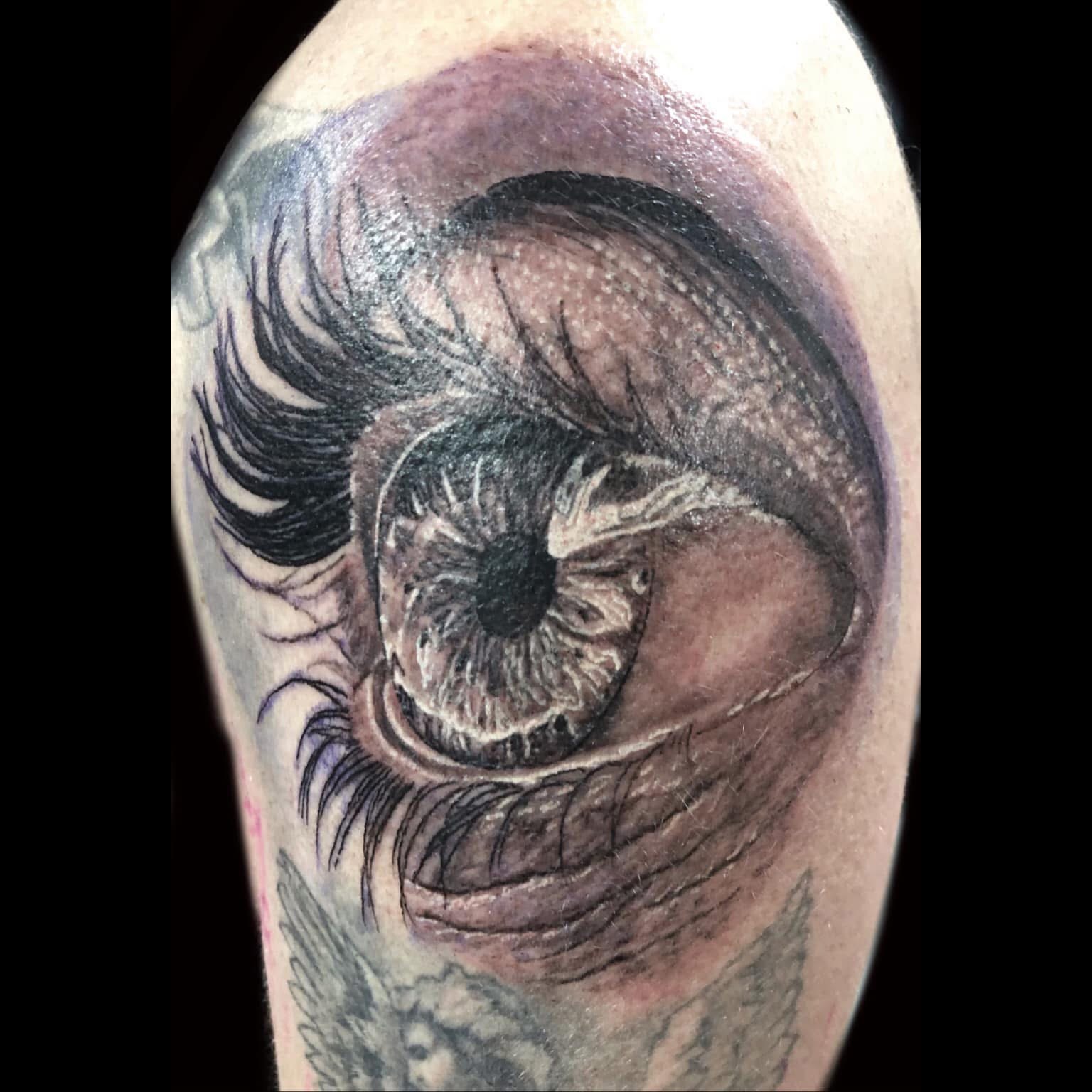 Black and grey hyper-realistic human eye with detailed iris and lashes on upper arm by Jin