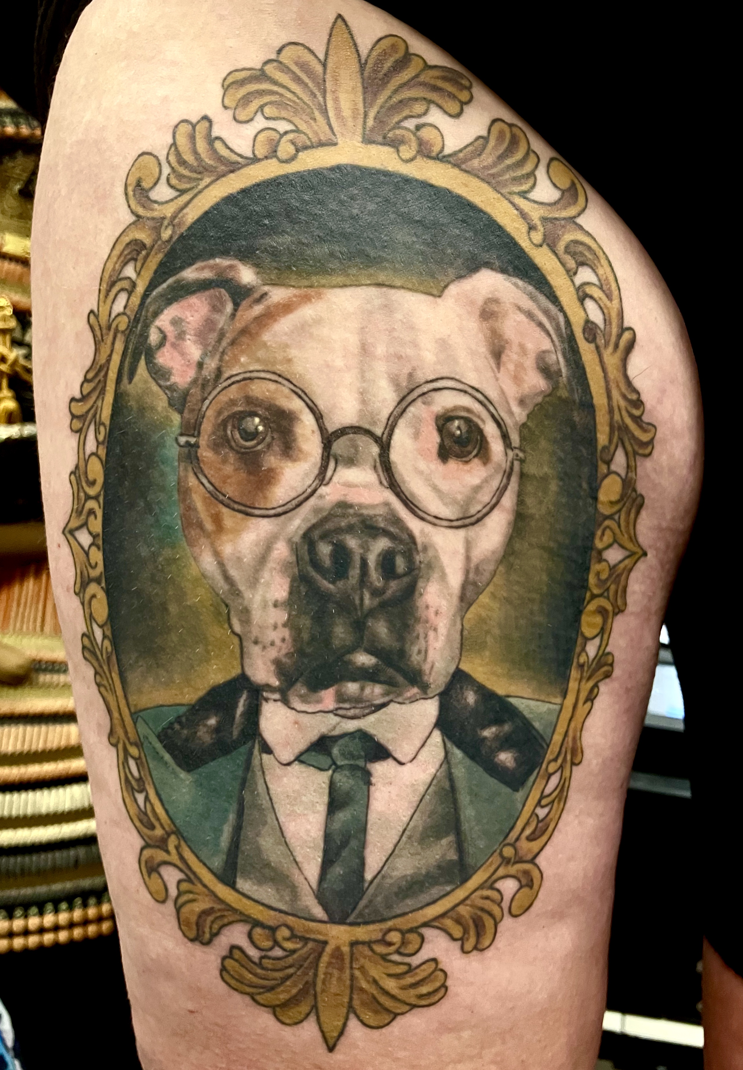 Realistic dog portrait in suit and glasses in ornate gold frame on thigh by Ray